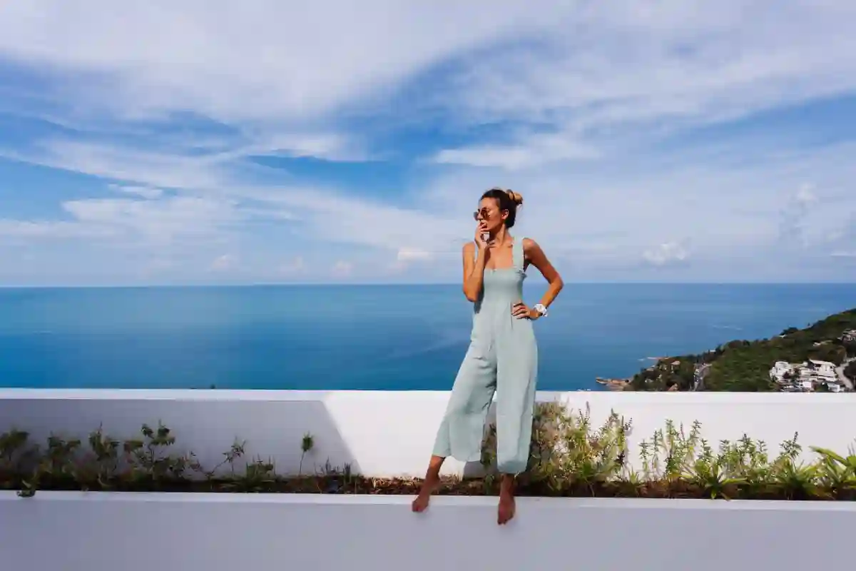 Fit tanned Caucasian woman in a light blue mint-colored overall posing on a terrace balcony, showcasing modern, casual chic style. Fit tanned Caucasian woman in a light blue mint-colored overall posing on a terrace balcony, showcasing modern, casual chic style.