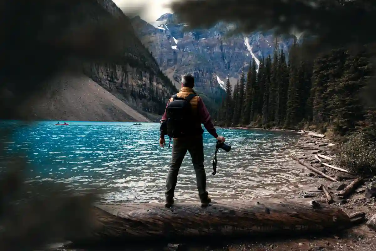 A man standing near a calm body of water in Alberta, Canada, holding a camera, ready to capture the scenery. A man standing near a calm body of water in Alberta, Canada, holding a camera, ready to capture the scenery.