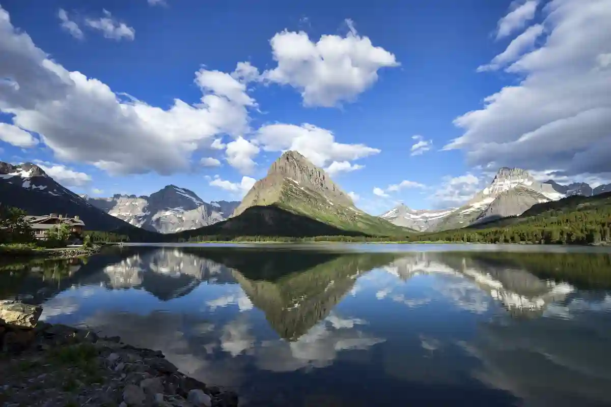 The serene waters of Swiftcurrent Lake reflecting the towering peaks of Glacier National Park, surrounded by lush greenery and wildflowers. The serene waters of Swiftcurrent Lake reflecting the towering peaks of Glacier National Park, surrounded by lush greenery and wildflowers.