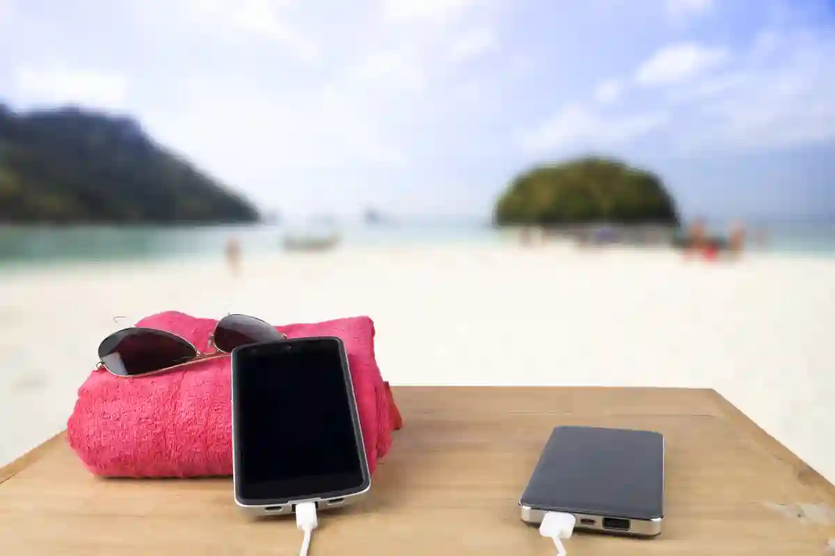 A red tower beside sunglasses, with a mobile phone charging via a power bank on a wooden table. A red tower beside sunglasses, with a mobile phone charging via a power bank on a wooden table.
