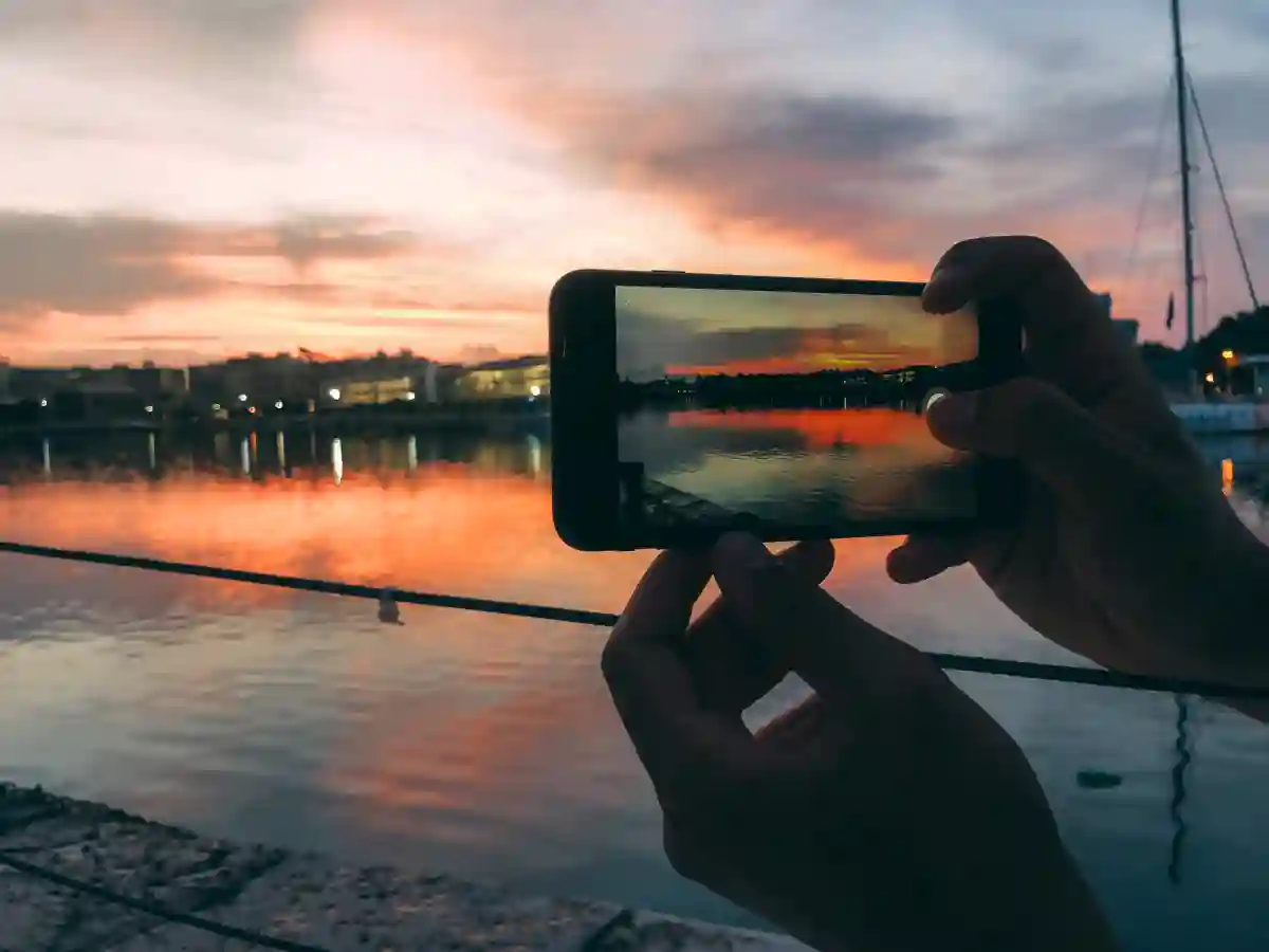 A man’s hands holding a mobile phone, snapping a photo of a vibrant sunset over the horizon. A man’s hands holding a mobile phone, snapping a photo of a vibrant sunset over the horizon.