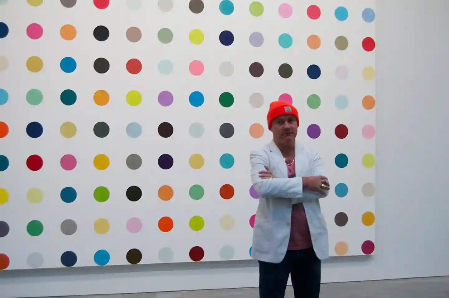 The image features British contemporary artist Damien Hirst posing in front of one of his signature "Spot Paintings". The image features British contemporary artist Damien Hirst posing in front of one of his signature "Spot Paintings".