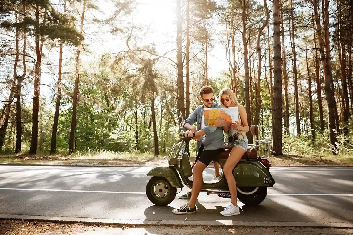 This image is a stock photo of a young couple on a road trip with a scooter, stopped on the side of a road in a wooded area, and looking at a map. This image is a stock photo of a young couple on a road trip with a scooter, stopped on the side of a road in a wooded area, and looking at a map.